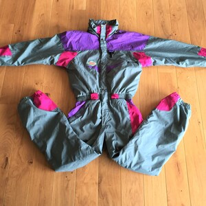 May include: A gray ski suit with pink and purple accents. The suit has a zipper closure and a hood.