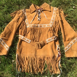 Puede incluir: Una camisa con flecos de ante color canela con cuello, mangas largas y cintur&oacute;n. La camisa presenta costuras decorativas blancas y flecos a lo largo de las mangas, el pecho y el dobladillo. La camisa se exhibe sobre hierba verde.