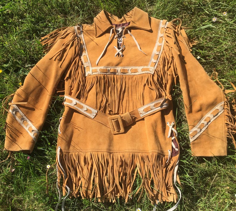 Puede incluir: Una camisa con flecos de ante color canela con cuello, mangas largas y cintur&oacute;n. La camisa presenta costuras decorativas blancas y flecos a lo largo de las mangas, el pecho y el dobladillo. La camisa se exhibe sobre hierba verde.