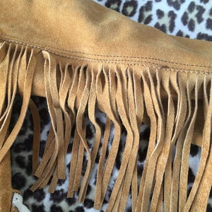 Puede incluir: Primer plano de una chaqueta de ante marr&oacute;n con flecos largos. Se ve el borde de la chaqueta, mostrando las costuras y la textura del ante. El fondo es un estampado de leopardo.