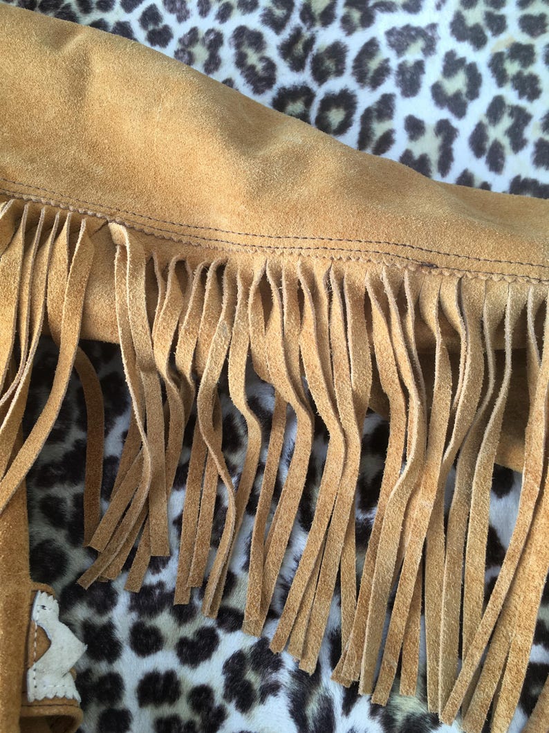 Puede incluir: Primer plano de una chaqueta de ante marr&oacute;n con flecos largos. Se ve el borde de la chaqueta, mostrando las costuras y la textura del ante. El fondo es un estampado de leopardo.