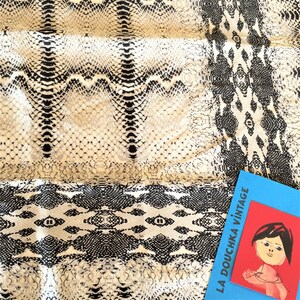 Può includere: Un tessuto bianco e nero con un motivo a pelle di serpente. Il tessuto ha un bordo con un motivo ripetitivo. Il tessuto è etichettato "LA DOUCHKA vintage".