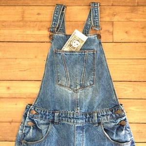 Könnte beinhalten: Hellblaue Jeans-Latzhose mit verstellbaren Trägern und einer Vordertasche. Die Latzhose hat ein klassisches Design mit Metallbeschlägen und eine Vintage-Ästhetik. Eine kleine Karte mit dem Text "LA DOUCHKA VINTAGE" befindet sich in der Vordertasche.