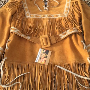 Puede incluir: Chaqueta de ante color mostaza con flecos largos. La chaqueta presenta un cuello, un cintur&oacute;n con hebilla cuadrada y cordones blancos. La chaqueta tiene ribetes blancos y una etiqueta que dice "La Douchka Vintage".