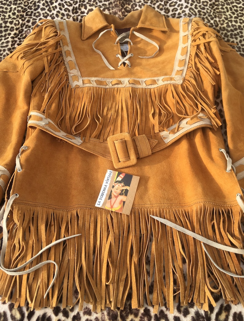 Puede incluir: Chaqueta de ante color mostaza con flecos largos. La chaqueta presenta un cuello, un cintur&oacute;n con hebilla cuadrada y cordones blancos. La chaqueta tiene ribetes blancos y una etiqueta que dice "La Douchka Vintage".
