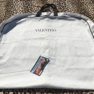 May include: A white garment bag with a black handle and trim. The word "VALENTINO" is printed in black across the top. A small tag with the text "LA DOUCHKA VINTAGE" and an illustration is attached to the bag.