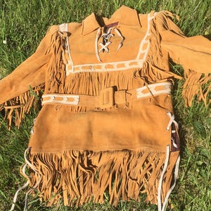 Puede incluir: Una camisa de ante marr&oacute;n con mangas largas y flecos. La camisa presenta un cuello, un cintur&oacute;n y detalles decorativos blancos. Los flecos est&aacute;n a lo largo de las mangas, el pecho y el dobladillo inferior.