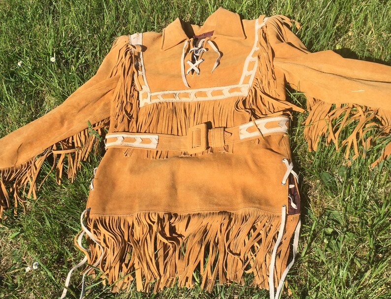 Puede incluir: Una camisa de ante marr&oacute;n con mangas largas y flecos. La camisa presenta un cuello, un cintur&oacute;n y detalles decorativos blancos. Los flecos est&aacute;n a lo largo de las mangas, el pecho y el dobladillo inferior.