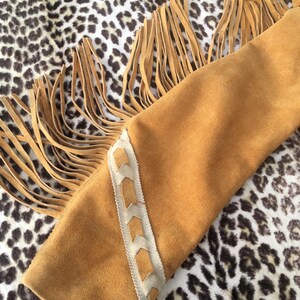 Puede incluir: Primer plano de una manga de chaqueta de ante marr&oacute;n con detalles de flecos largos. La manga tiene un patr&oacute;n decorativo cosido en color marr&oacute;n claro a lo largo del borde. El fondo es un estampado de leopardo.