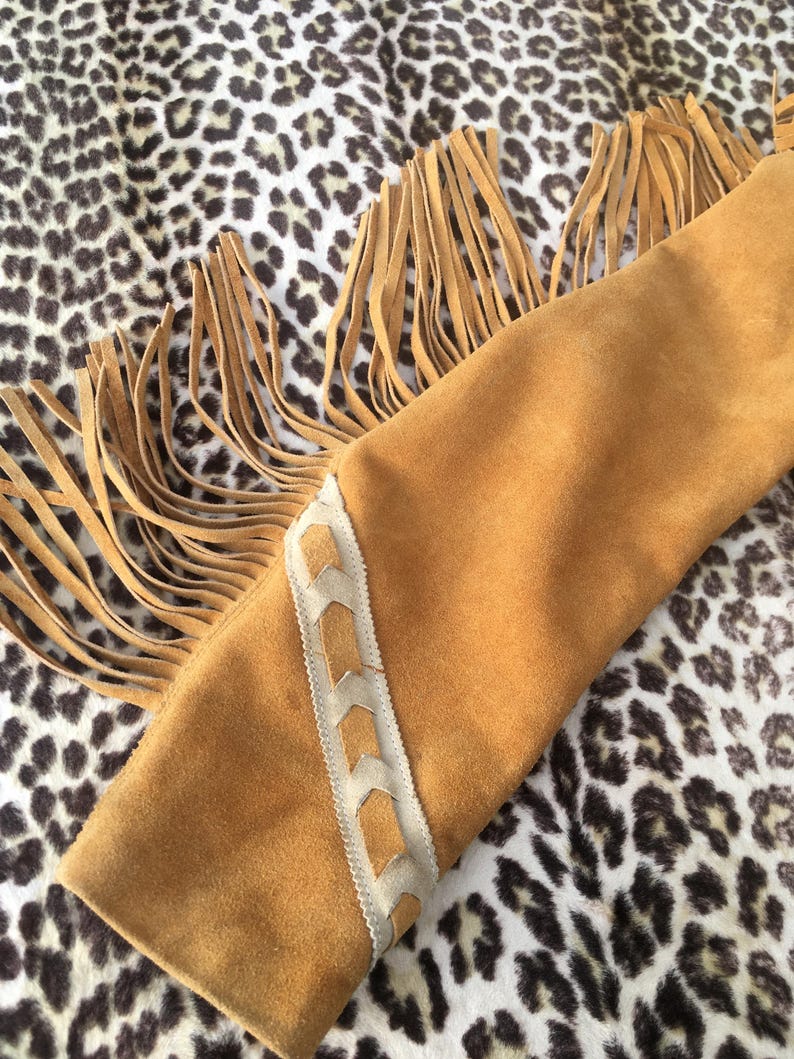 Puede incluir: Primer plano de una manga de chaqueta de ante marr&oacute;n con detalles de flecos largos. La manga tiene un patr&oacute;n decorativo cosido en color marr&oacute;n claro a lo largo del borde. El fondo es un estampado de leopardo.