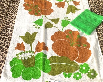 May include: White vintage bath towel with large orange, green, and brown floral pattern. A small green washcloth is next to the towel. A tag with the text "LA DOUCHKA VINTAGE" is in the bottom corner.