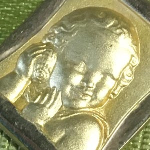 以下が含まれることがあります： 金色のペンダント。ケルビムの姿が詳細にレリーフされています。その姿は小さな物、おそらく花か信仰の象徴を持っています。ペンダントは長方形で、暗い縁取りがあり、緑色の背景に配置されています。