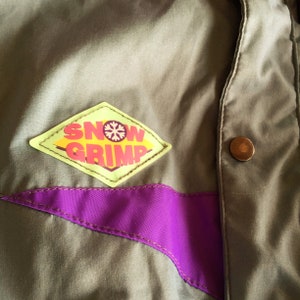 May include: A green jacket with a purple stripe and a yellow diamond-shaped patch that says "SNOW GRIMP" in red letters with a snowflake in the middle.
