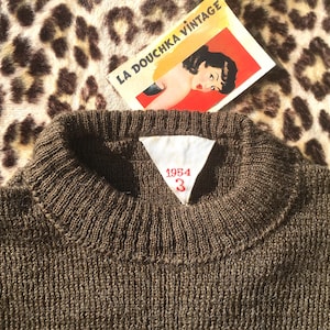 Peut inclure: Un pull en tricot marron avec un col côtelé. Une étiquette blanche avec les chiffres "1954" et "3" en rouge est cousue dans le col. L'étiquette vintage "LA DOUCHKA VINTAGE" est visible en arrière-plan.