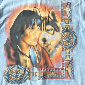 May include: A vintage t-shirt with a blue background featuring a Native American woman with a wolf. The image is surrounded by a beaded border with traditional Native American designs. The text "Authentic Traditional Indian Art" is at the bottom of the shirt.
