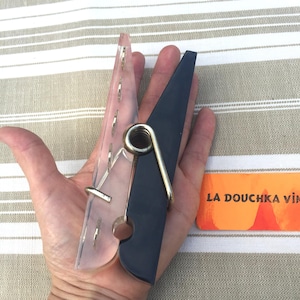 Pode incluir: Um grande prendedor de roupa decorativo com um lado de plástico transparente texturizado e um lado preto, conectados por uma mola de metal prateado. O prendedor de roupa é segurado em uma mão, com um pequeno cartão laranja que diz "LA DOUCHKA VIN" no fundo.