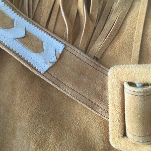 Puede incluir: Primer plano de un bolso de ante marr&oacute;n con largas flecos. El bolso presenta una correa decorativa blanca y marr&oacute;n con un dise&ntilde;o geom&eacute;trico. Se puede ver un peque&ntilde;o bolsillo y una hebilla.