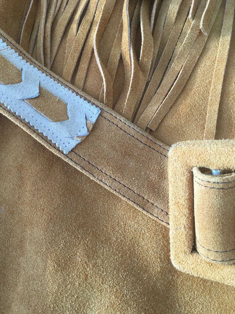 Puede incluir: Primer plano de un bolso de ante marr&oacute;n con largas flecos. El bolso presenta una correa decorativa blanca y marr&oacute;n con un dise&ntilde;o geom&eacute;trico. Se puede ver un peque&ntilde;o bolsillo y una hebilla.