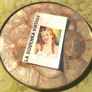 May include: A round decorative tray with a gold rim and a mother-of-pearl mosaic surface. A vintage postcard with the text "LA DOUCHKA VINTAGE" and a retro illustration of a woman is placed on the tray.