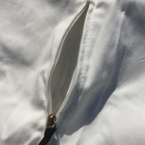 May include: Close-up of white fabric with a zipper and a gold-colored grommet. The zipper is gold-colored with a black pull tab. The fabric has a subtle texture.