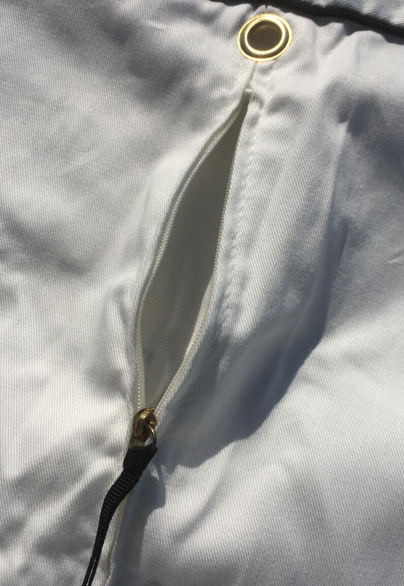 May include: Close-up of white fabric with a zipper and a gold-colored grommet. The zipper is gold-colored with a black pull tab. The fabric has a subtle texture.