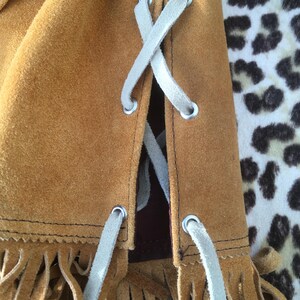 Puede incluir: Primer plano de un bolso de ante marr&oacute;n con cordones blancos en forma de X. El bolso tiene flecos en el borde inferior. El fondo es un estampado de leopardo.