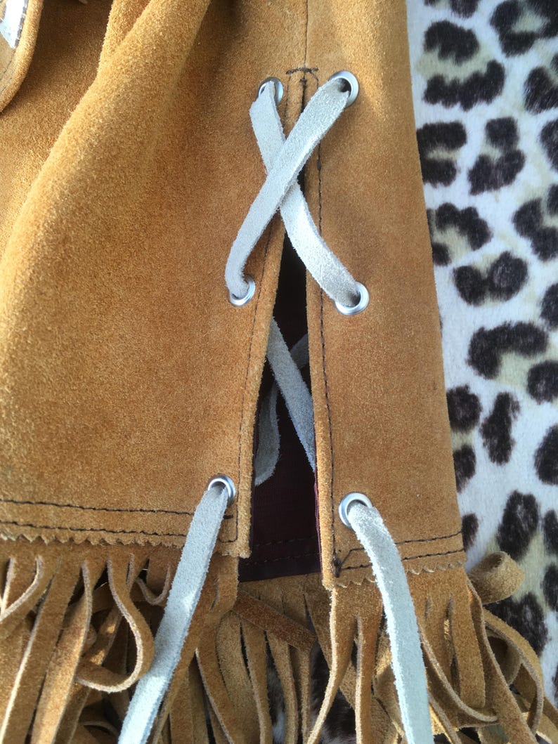 Puede incluir: Primer plano de un bolso de ante marr&oacute;n con cordones blancos en forma de X. El bolso tiene flecos en el borde inferior. El fondo es un estampado de leopardo.