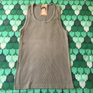 May include: A ribbed, olive-green tank top with a scoop neck. The top has a tag attached with the text "LA DOUCHE VINTAGE" and an illustration of a woman. The tank top is displayed on a green and white patterned background.
