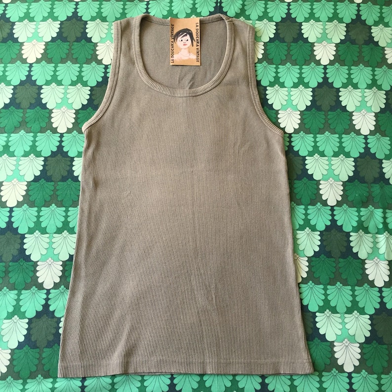May include: A ribbed, olive-green tank top with a scoop neck. The top has a tag attached with the text "LA DOUCHE VINTAGE" and an illustration of a woman. The tank top is displayed on a green and white patterned background.
