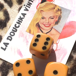 May include: Three orange dice with black dots are displayed. A vintage-style image of a winking woman is in the background, along with a card that reads "LA DOUCHKA VINTAGE". The dice are a warm, inviting color.