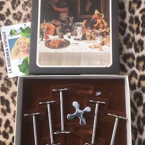 Puede incluir: Un juego de cubiertos vintage Jean Couzon, con una caja con una pintura de una escena de comedor y un juego de piezas para servir de metal plateado. La caja también contiene una tarjeta vintage con el texto "La Douchka Vintage".