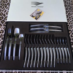 May include: A Pradel France cutlery set in a white box with a brown interior tray. The set includes silver-toned knives, forks, and spoons with curved handles. A small square card with a vintage illustration and the text LA DOUCHKA VINTAGE is placed on the inside lid.