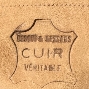 May include: Close-up of a tan leather surface with an embossed emblem. The emblem reads "DESSUS & DESSOUS CUIR VERITABLE" in a stylized font, indicating genuine leather. The leather has a textured appearance.