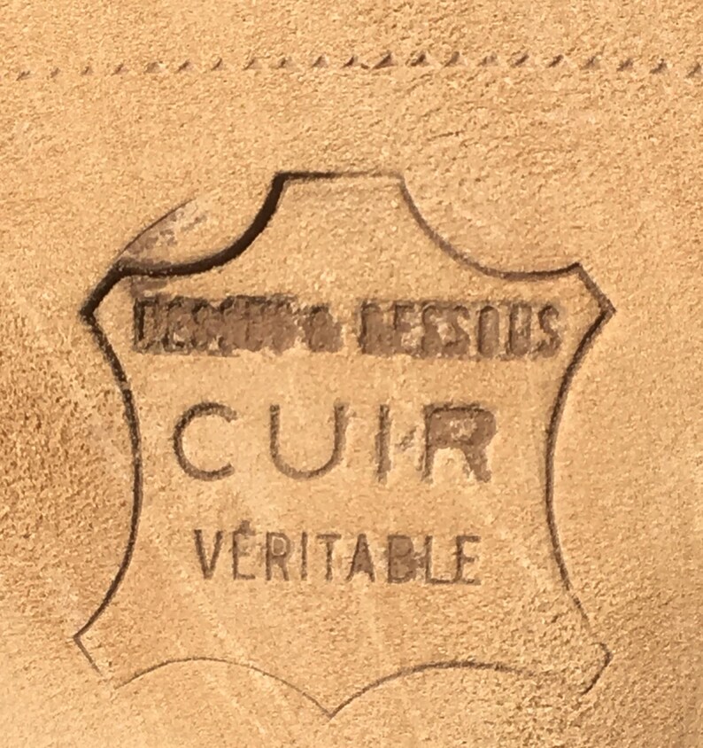 May include: Close-up of a tan leather surface with an embossed emblem. The emblem reads "DESSUS & DESSOUS CUIR VERITABLE" in a stylized font, indicating genuine leather. The leather has a textured appearance.
