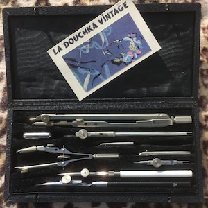 Könnte beinhalten: Ein Vintage-Zeichensatz in einem schwarzen Koffer. Das Set enthält verschiedene silberfarbene Zirkel, Teiler und andere Zeichenwerkzeuge. Eine Karte mit dem Text "LA DOUCHKA VINTAGE" und einer Illustration befindet sich im Inneren des Koffers.