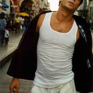 May include: A man in a white tank top and light-colored pants stands on a city street. He wears a black jacket draped over his shoulders. The background shows a bustling street scene with buildings and people.