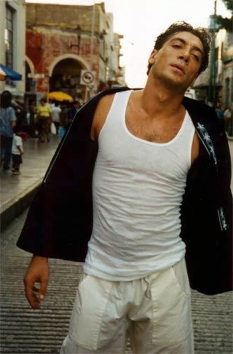 May include: A man in a white tank top and light-colored pants stands on a city street. He wears a black jacket draped over his shoulders. The background shows a bustling street scene with buildings and people.