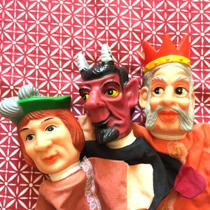 Puede incluir: Tres marionetas de mano vintage: un bufón con un sombrero verde, un diablo rojo con cuernos y un rey con una corona. Las marionetas están hechas de plástico y tela, con rasgos faciales detallados. El fondo es una tela estampada en rojo y blanco.