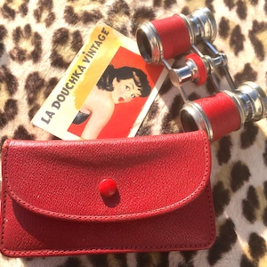 May include: Vintage red opera glasses with silver accents and a matching red leather case. A vintage advertisement card with the text "LA DOUCHKA VINTAGE" is also present. The items are displayed on a leopard print surface.