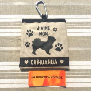 以下が含まれることがあります： 黒いチワワのシルエット、足跡、そして「J'AIME MON CHIHUAHUA」の文字がプリントされた、黒い縁取りのあるベージュ色のキャンバスバッグ。シルバーのカラビナクリップ付き。バッグの下には、オレンジと赤色の小さなカードがあります。
