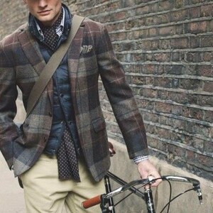 May include: A man wearing a brown and black plaid jacket, a navy blue vest, a white shirt with a black and white patterned tie, khaki pants, and a brown newsboy cap. He is holding a black bicycle with a brown leather handlebar grip.