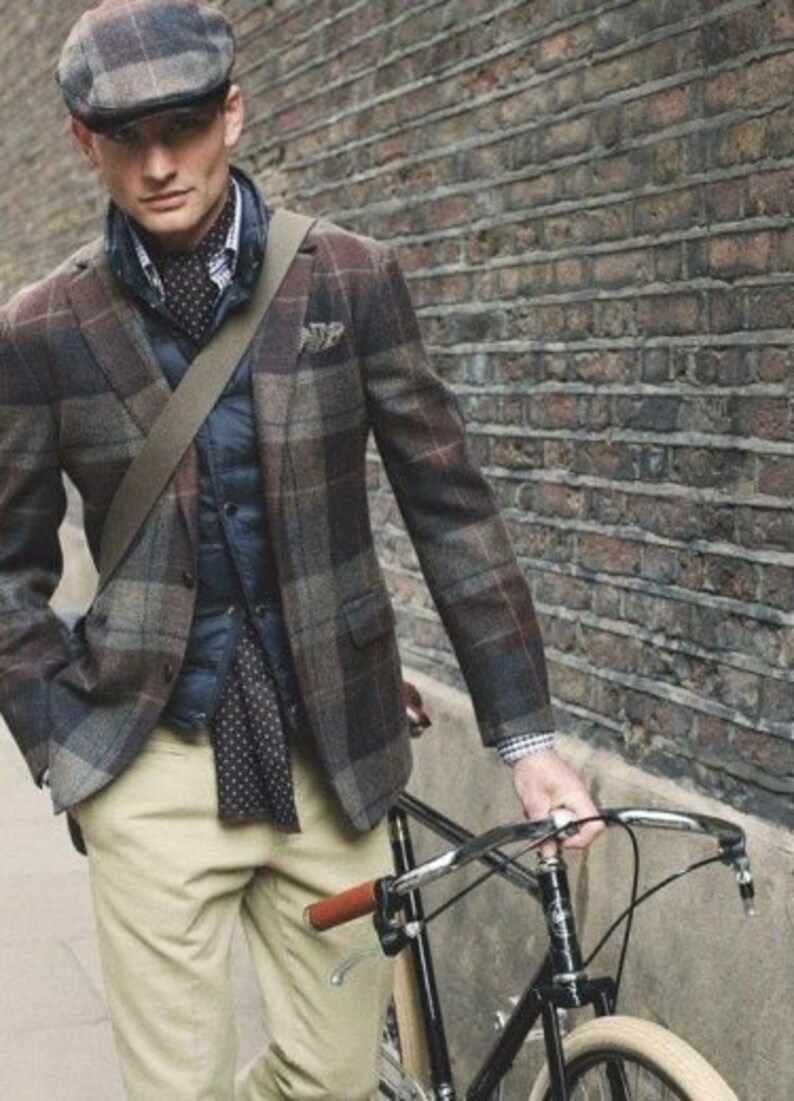 May include: A man wearing a brown and black plaid jacket, a navy blue vest, a white shirt with a black and white patterned tie, khaki pants, and a brown newsboy cap. He is holding a black bicycle with a brown leather handlebar grip.
