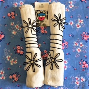 May include: Two white napkins with black metal napkin rings in the shape of a sun. The rings are on a blue and white floral tablecloth. A card with the text "LA DOUCHKA VINTAGE" is visible in the image.