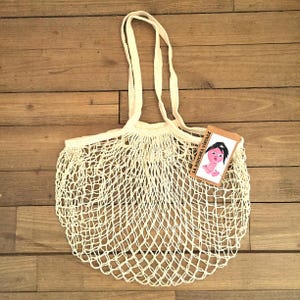 Op de afbeelding: Een witte netten tas met een bruin label waarop "LA DOUCHKA VINTAGE" staat en een cartoontekening van een vrouwen gezicht. De tas heeft twee lange handvatten.