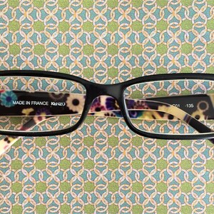 Puede incluir: Gafas con montura negra con un patrón floral colorido en el interior de las lentes. El texto "MADE IN FRANCE KENZO" es visible en la lente izquierda.