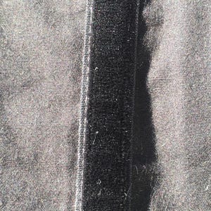May include: Close-up of a black hook and loop fastener strip sewn onto a gray fabric. The hook and loop strip is centered, with the fabric visible on either side. The fabric has a textured appearance.