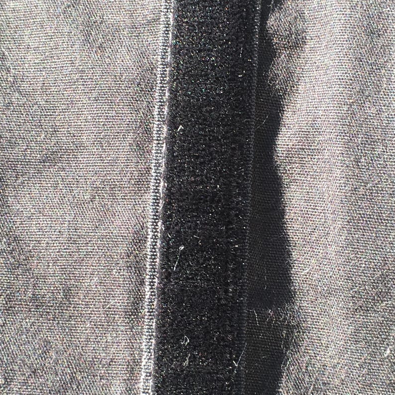 May include: Close-up of a black hook and loop fastener strip sewn onto a gray fabric. The hook and loop strip is centered, with the fabric visible on either side. The fabric has a textured appearance.