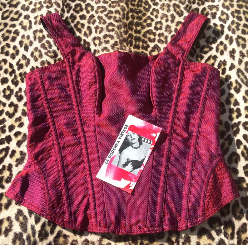 May include: A magenta-colored corset top with shoulder straps. The top has vertical seams and a textured fabric. A tag with a vintage image and the text "LA DOUCHKA VINTAGE" is attached to the garment. The background is a leopard print.