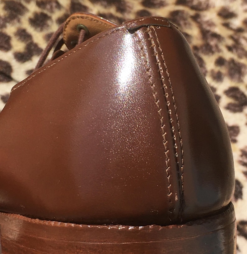 May include: Close-up of a brown leather shoe, showcasing the polished surface and detailed stitching along the side. The shoe's heel and sole are visible, highlighting the craftsmanship of the footwear.