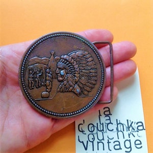 Peut inclure: Une boucle de ceinture vintage en cuivre vieilli représentant un chef amérindien de profil portant une coiffe à plumes. La boucle représente une scène avec un tipi et des personnages en arrière-plan.